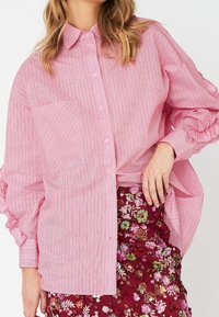 Femme portant une chemise oversize à rayures roses et blanches avec des manches à volants et une jupe fleurie rouge ornée de sequins.