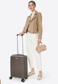 Hartschalenkoffer in Dunkelbraun mit horizontalen Rillen, silbernem Griff und vier Rädern. Kombiniert mit einer beigen Lederjacke, cremfarbenen Hosen und einer kleinen Handtasche.