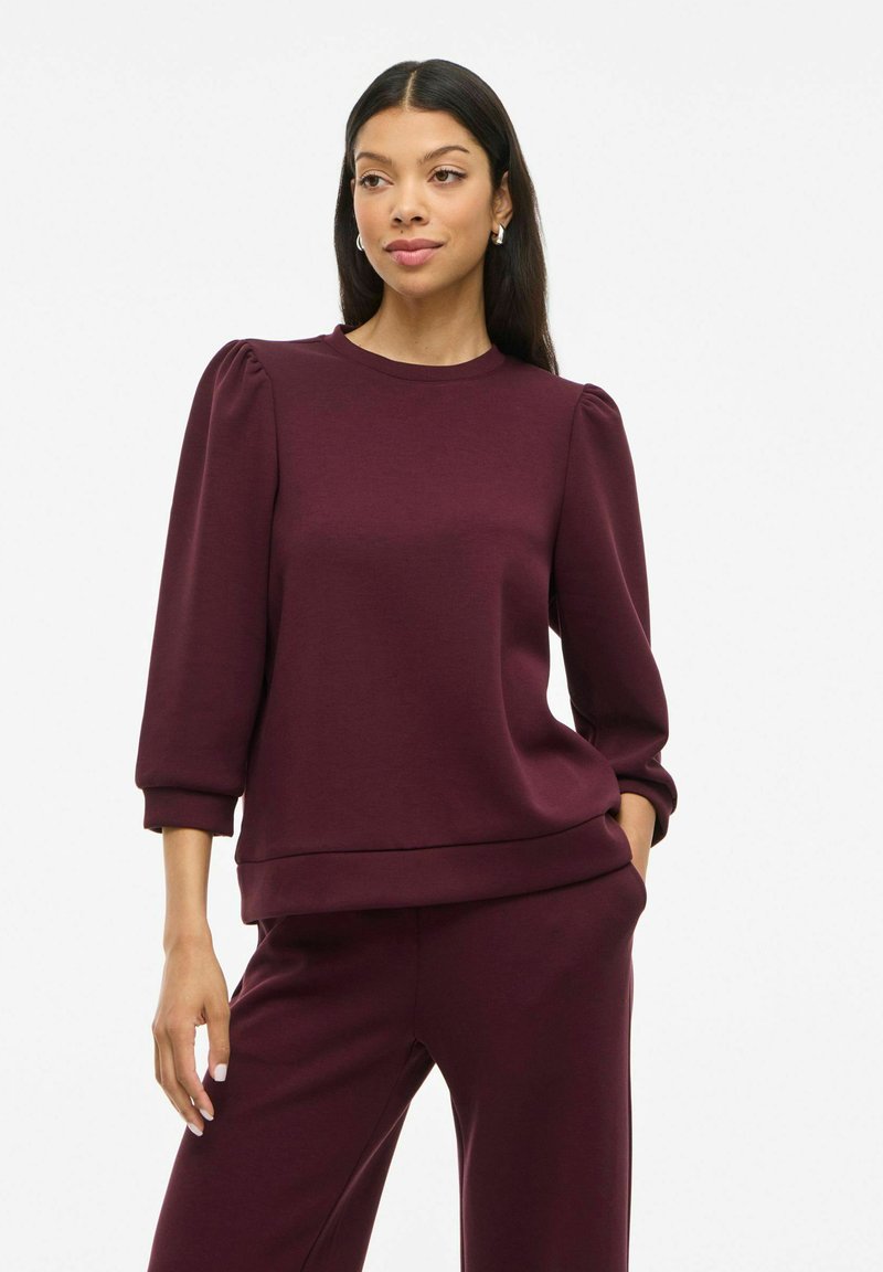 Bordeaux genser med trekvart puffermer, rund hals og et mykt, teksturert stoff. Løs passform og ribbestrikket kant.