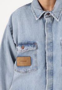 Gai zila džinsa jaka ar klasisku apkakli, divām priekšējo kabatām, brūnu ādas etiķeti ar uzrakstu "H2OFagerholt" un redzamām šuvju detaļām.