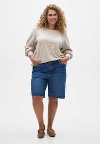 Beige knitted sweater with a diamond pattern, paired with blue denim shorts. Model wears brown flats. Neutral background.
