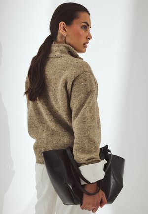 Beige gebreide trui met hoge kraag, losse fit en gestructureerde spikkels; zwarte handtas met korte handvatten. Witte manchetten zichtbaar.