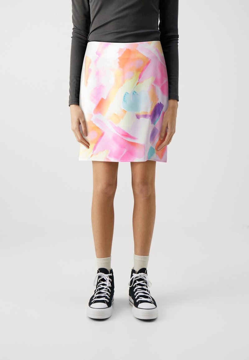 Femme portant une jupe à imprimé abstrait pastel multicolore, un haut noir à manches longues, des chaussettes blanches et des baskets montantes noires, debout sur un fond blanc.