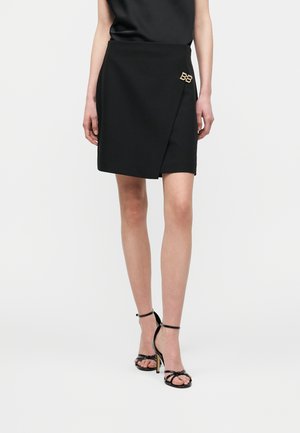 Femme portant une jupe portefeuille noire avec un détail du logo BB doré, associée à des sandales à talons hauts à bride cheville noires, sur un fond uni.