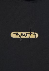 Tela de prenda negra con un logo bordado en oro sobre un fondo rectangular liso y letras cursivas estilizadas.