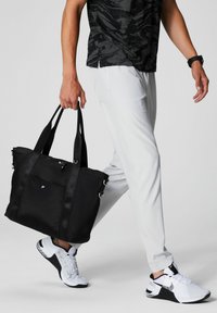 Black nylon tote bag with dual straps, zip closure, front pocket, and accent hardware, being carried by a person in light pants and sneakers.