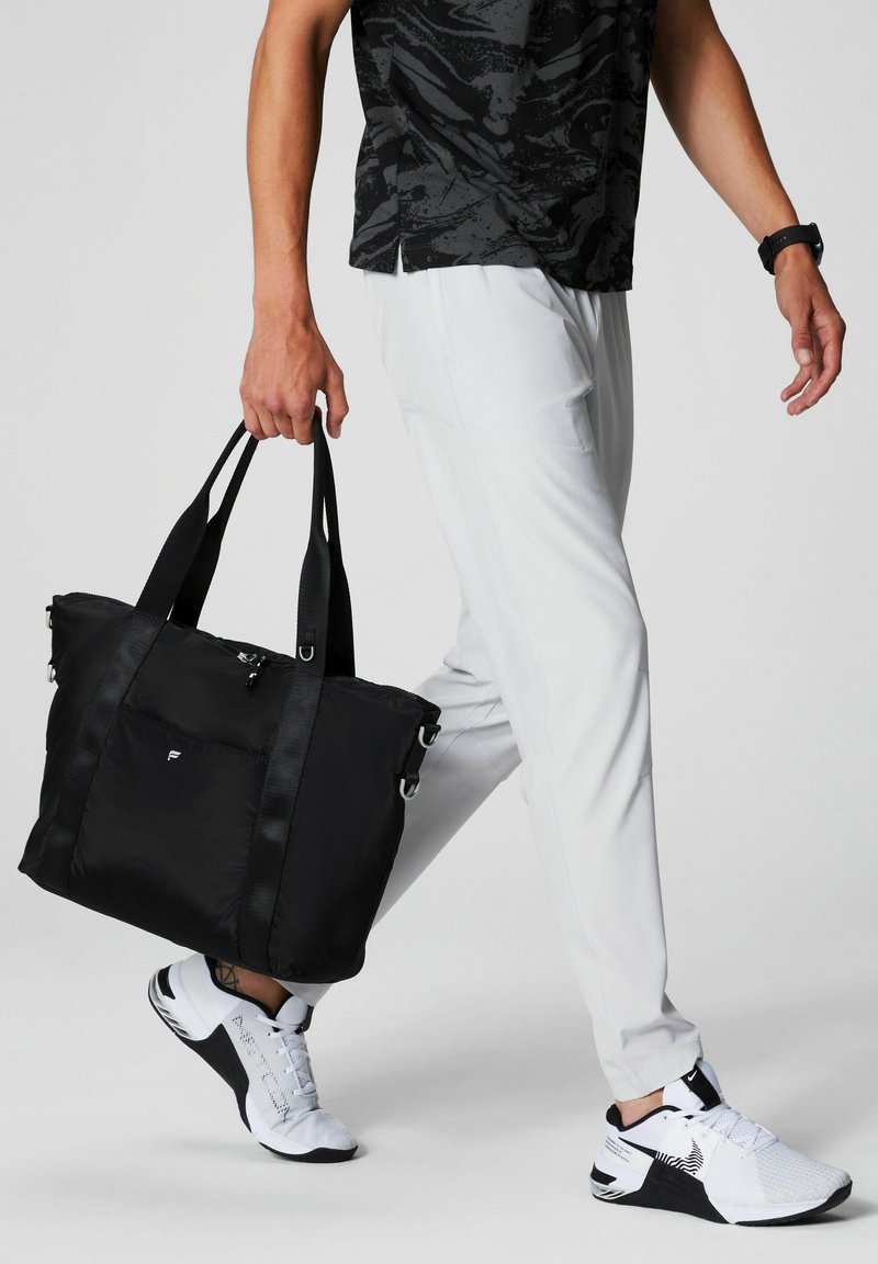 Black nylon tote bag with dual straps, zip closure, front pocket, and accent hardware, being carried by a person in light pants and sneakers.