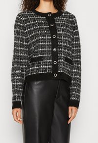 Cardigan a scacchi bianco e nero con collo rotondo, bottoni frontali, due tasche, tessuto texturizzato e polsini a costine, indossato su pantaloni neri.