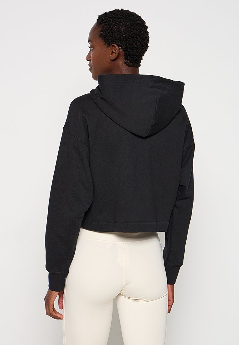 Persona che indossa un hoodie corto nero con il cappuccio abbassato e leggings beige a vita alta, in piedi e di spalle su uno sfondo bianco semplice.