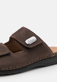 Braune Leder-Sandale mit verstellbarem Riemen, genähten Details, einem metallischen Logo-Akzent und einer gepolsterten Innensohle auf einer schwarzen Gummisohle.