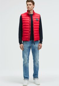Rote Steppweste mit hohem Kragen und Frontreißverschluss, kombiniert mit dunklen langen Ärmeln und hellblauen Jeans, abgerundet mit weißen Turnschuhen.