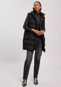 Czarna ocieplana kamizelka z kapturem, z pełnym zamkiem, srebrnymi akcentami i bocznymi kieszeniami, noszona na czarnym długim topie i szarych dżinsach.