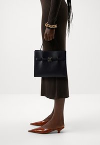 Woman wearing ribbed brown dress, brown kitten heel shoes, and gold chain bracelet holding black DK handbag by her side.