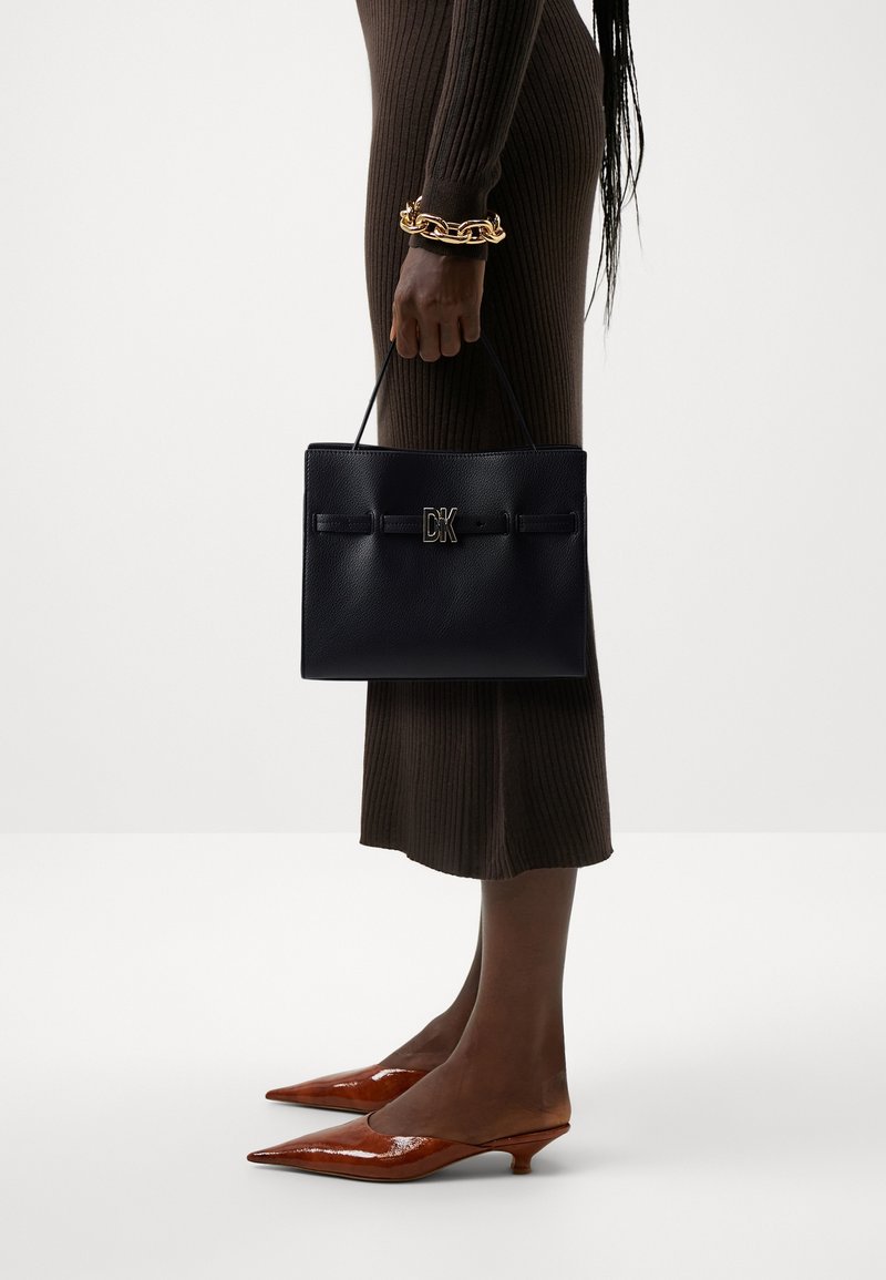 Woman wearing ribbed brown dress, brown kitten heel shoes, and gold chain bracelet holding black DK handbag by her side.