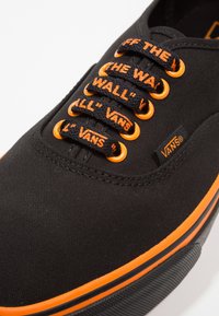 Baskets noires en toile avec des accents orangés, texte "VANS" et "OFF THE WALL" sur les lacets, et une semelle en caoutchouc avec une bande contrastante.