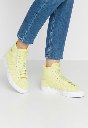 Personne portant des baskets montantes Adidas jaune clair avec semelles blanches et jeans courts bleus, debout sur un sol blanc.