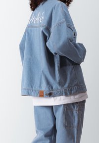 Personne portant une veste et un pantalon en jean bleu clair avec un texte brodé blanc au dos de la veste et sur la poche du pantalon, superposés sur une chemise blanche.