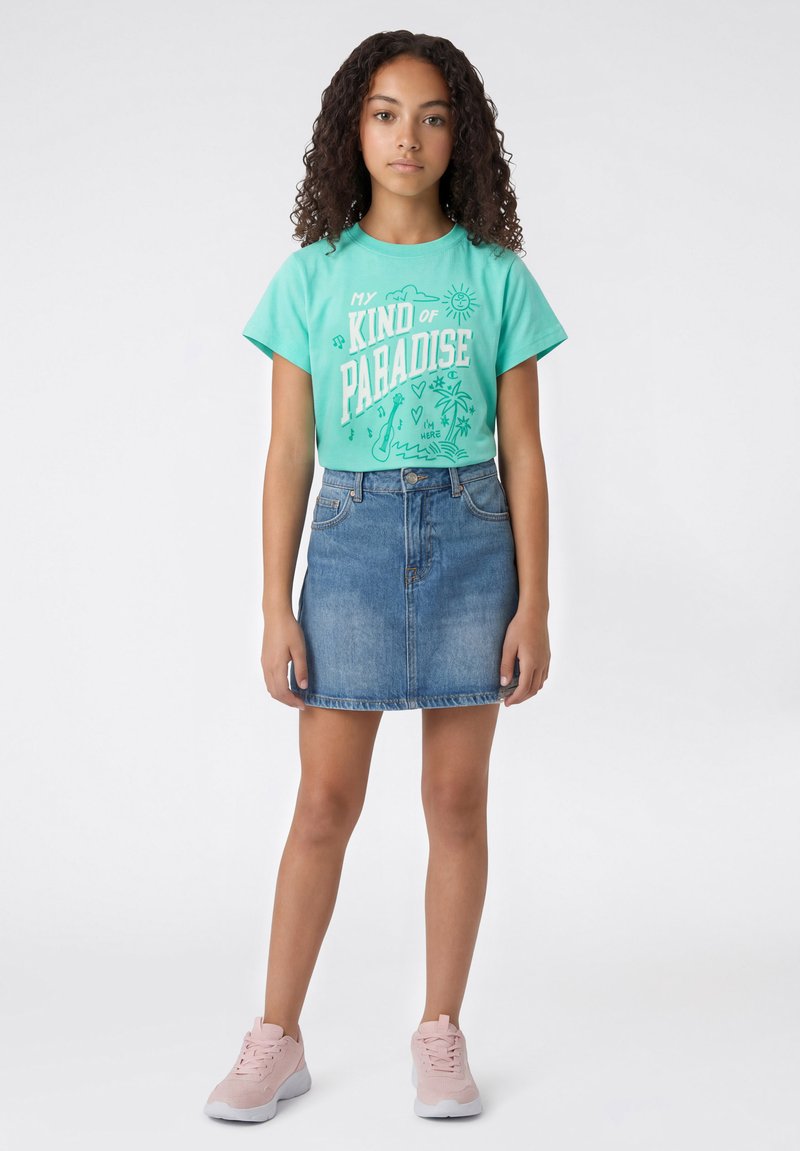Jeune fille aux cheveux bouclés portant un t-shirt turquoise "My Kind of Paradise", une jupe en denim et des baskets roses, debout devant un fond blanc.