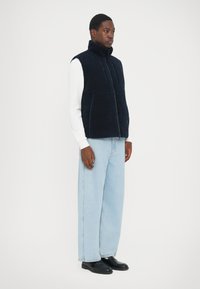 Gilet en polaire marine avec fermeture éclair, col montant et deux poches avant, porté sur un haut blanc et associé à un jean évasé bleu clair.