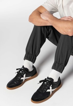 Chaussures de sport noires avec une bande diagonale blanche, en cuir et en daim, semelle en caoutchouc marron, portées avec un pantalon noir et des chaussettes blanches.