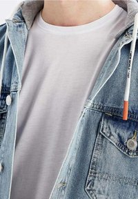 Kangashame denim, jossa on harmaa huppu, sisältäen vaaleansinistä kangasta, metallipainikkeita ja näkyvää ommelta. Alle näkyy tavallinen valkoinen t-paita.