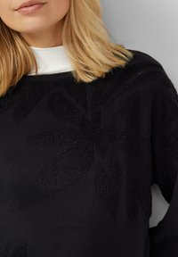 Maglione nero con motivo floreale a rilievo, scollo rotondo e maniche lunghe. Il tessuto sembra morbido e leggermente vellutato. Camicia bianca visibile sotto.