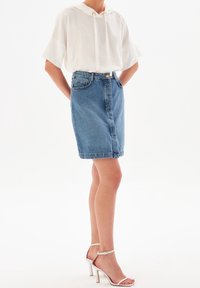 Kvinna iklädd en vit kortärmad blus, knälång blå denimkjol och vita högklackade sandaler mot en vit bakgrund.