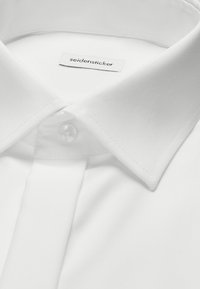 Biała koszula z klasycznym spiczastym kołnierzykiem, zapinana na pojedynczy biały guzik, z metką "seidensticker" od wewnątrz kołnierzyka.