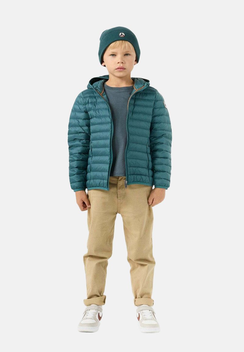 Petrolblauwe donsjas met capuchon en ritssluiting aan de voorkant, gedragen over een grijs shirt. Beige broek en witte sneakers met rode accenten. Gebreide petrolblauwe muts.