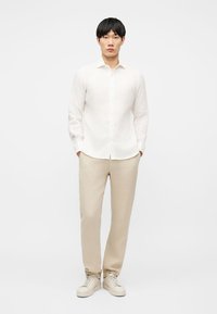 Άνδρας όρθιος με τα χέρια στις τσέπες, φορώντας λευκό πουκάμισο με κουμπιά, μπεζ παντελόνι και μπεζ αθλητικά παπούτσια, στο φόντο ενός απλού σκηνικού.