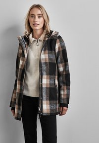 Cappotto a quadri in una miscela di lana nera, beige e bianca con cappuccio, chiusura a zip e tasche laterali. Indossato sopra un maglione crema e pantaloni scuri.