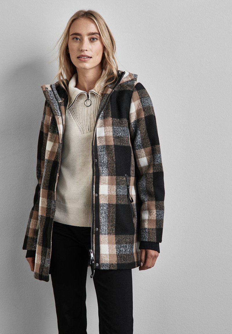 Cappotto a quadri in una miscela di lana nera, beige e bianca con cappuccio, chiusura a zip e tasche laterali. Indossato sopra un maglione crema e pantaloni scuri.