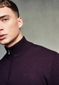Paarse gebreide pullover met een hoge ritssluiting, gekenmerkt door een gestructureerd patroon. De ritssluiting heeft een onderscheidende metalen trekker.