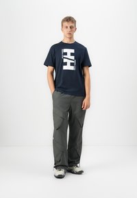 Junger Mann mit Händen in den Taschen, trägt ein navyblaues Helly Hansen Logo-T-Shirt, graue Cargo-Hosen und weiße Wanderschuhe vor einem schlichten weißen Hintergrund.