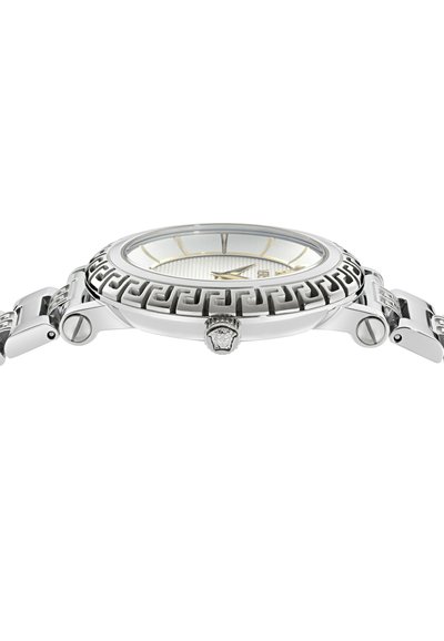 Reloj de pulsera plateado con bisel de patrón de clave griega, manecillas plateadas y doradas, y esfera blanca texturizada, mostrado en perfil lateral.