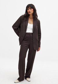 Costume marron ajusté avec une veste à un seul bouton et un pantalon à jambe droite. Porté sur un débardeur blanc, avec deux boutons et des poches.