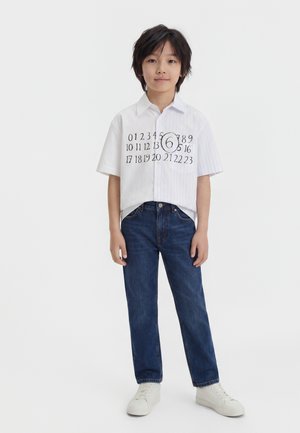Dieťa s čiernymi vlasmi oblečené v bielej krátkotretej košeli s potlačou kalendárnych čísel a tmavomodrých džínsoch stojace na bielom pozadí.