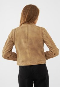 Brun läderjacka med en figurnära design, med ribbade axelaccenter och en mjuk yta. Bärs med svarta byxor, sedd bakifrån.