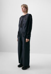 Zwart technisch outfit met een ritsoverslag met lange mouwen, zakken en brede broekspijpen. Gladde textuur, gestructureerd ontwerp en relaxte pasvorm.