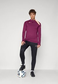 Paarse atletische top met lange mouwen, hoge kraag en open schouderdetail, gecombineerd met zwarte strak aansluitende broek en zwarte voetbalschoenen.