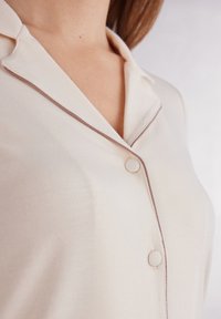 Ljus beige blus med krage, med bruna kantlinjer och runda knappar. Slät textur och skräddarsydd passform är tydlig.