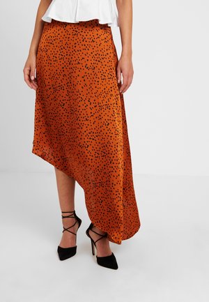 Femme portant une jupe asymétrique orange à taches noires, associée à des talons hauts noirs à brides et un haut blanc.