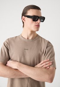 De grandes lunettes de soleil noires avec des montures angulaires, des verres sombres et des accents texturés sur les branches. Portées avec un T-shirt beige clair arborant un logo.