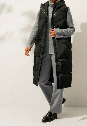 Gilet matelassé noir avec fermeture éclair et capuche, associé à un pull en maille gris clair et un pantalon gris ample, porté avec des mocassins noirs.