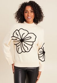 Maglione crema con scollo e polsini a coste. Presenta una grande ricamatura floreale nera sulla parte anteriore e sulle maniche. Abbinato a pantaloni in pelle nera.