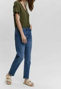 Femme portant un haut vert olive à manches courtes et boutonné, un jean taille haute bleu et des sandales beige à double bride sur fond uni.