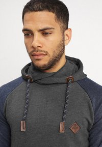 Sudadera gris con capucha y mangas raglán en color marino, cordones con detalles de cuero y un parche de logo en forma de diamante en el pecho. Textura de tejido suave.