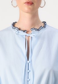 Blusa blu chiaro con colletto arricciato bianco e nero, scollatura a forma di chiave e bottoni frontali. Tessuto liscio con una consistenza morbida.