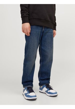 Person iført løse blå jeans, sort hættetrøje og hvide sneakers med blå detaljer, stående på et lyst gråt gulv.