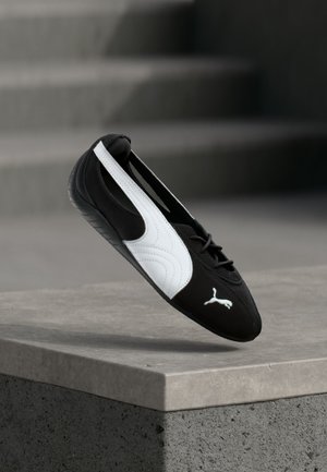 Sort-hvid Puma sneaker med snørebånd, svævende over en betonkant mod en sløret grå trappebaggrund.